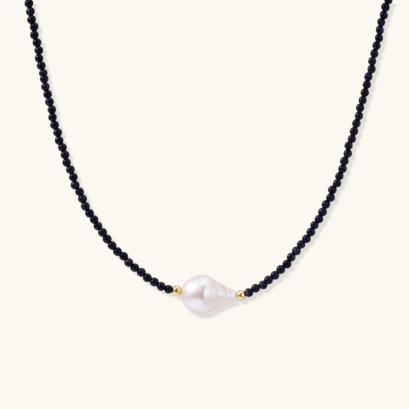 Delicate necklace with black blue sandstone beads and baroque pearl center on white background with gold accents.