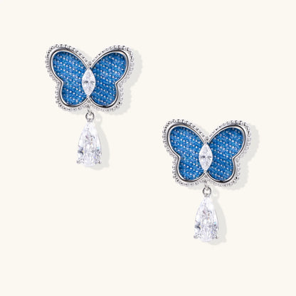 Blue denim butterfly earrings with silver frame and teardrop crystal dangles on cream background