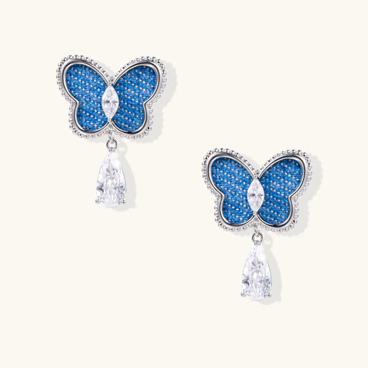 Blue denim butterfly earrings with silver frame and teardrop crystal dangles on cream background