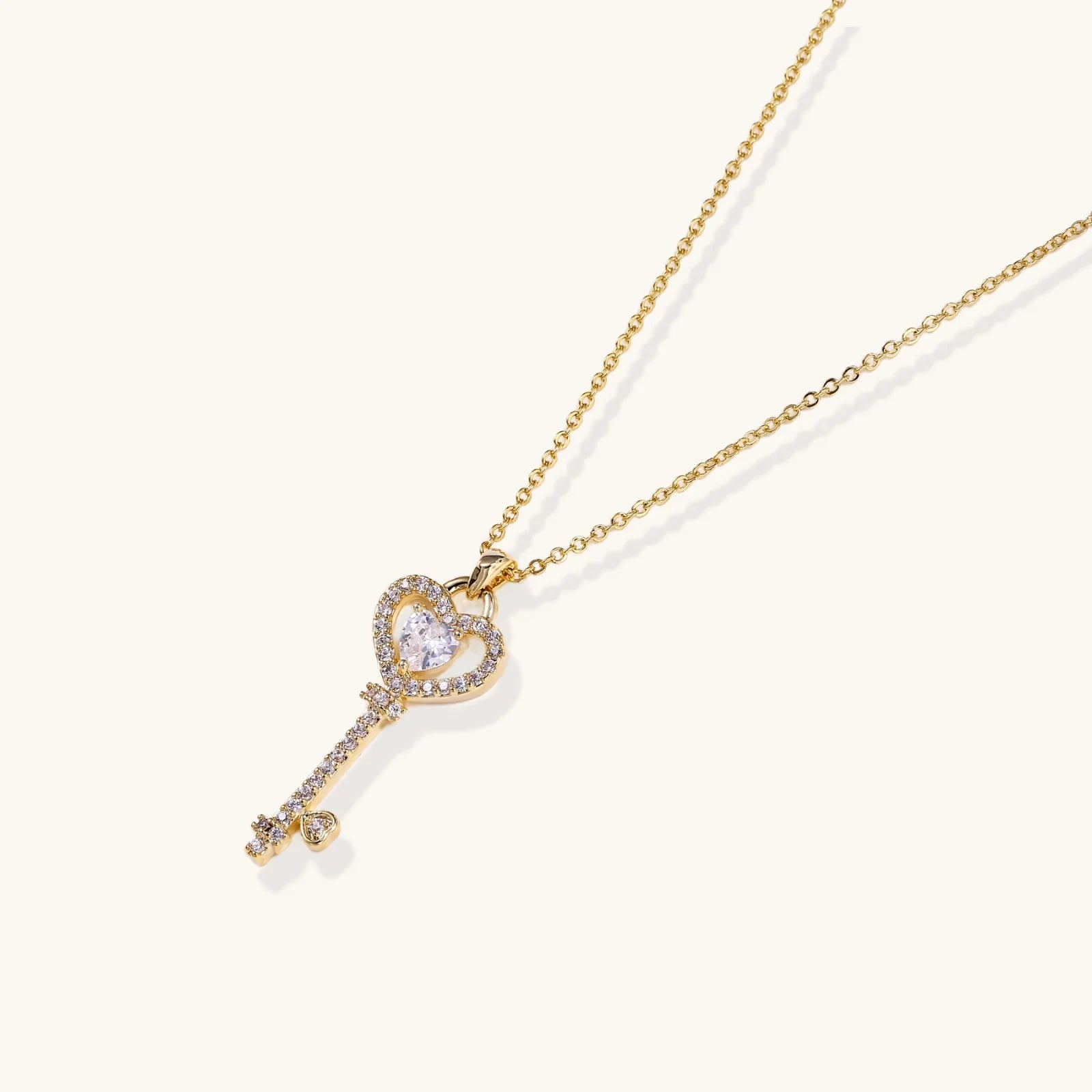 Delicate gold key pendant with heart-shaped bow and crystal pavé detailing on fine chain against cream background.