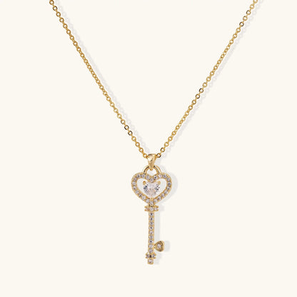 Delicate gold key pendant with heart-shaped bow and crystal pavé detailing on fine chain against cream background.